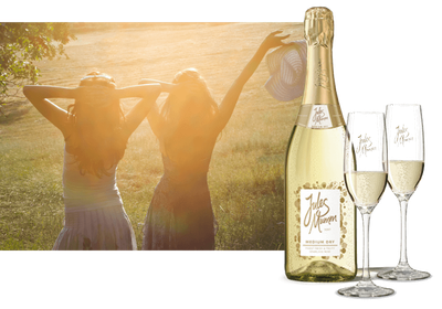 Eine Flasche Jules Mumm Sekt und zwei Gläser stehen im Vordergrund, während zwei Frauen im Sonnenlicht Freude ausstrahlen.