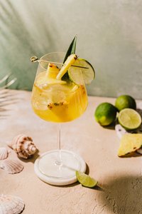 Ein erfrischender Cocktail in einem eleganten Glas, garniert mit Ananas und Limette, auf einem sandfarbenen Tisch.