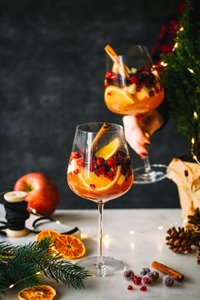 Auf einem Tisch stehen Gläser mit einem fruchtigen Getränk, umgeben von Tannenzweigen, getrockneten Orangen und Lichtern.
