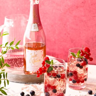Auf einem Tisch stehen zwei Gläser mit prickelndem Rosé, dekoriert mit Beeren und Minze, während eine Flasche im Hintergrund sichtbar ist.