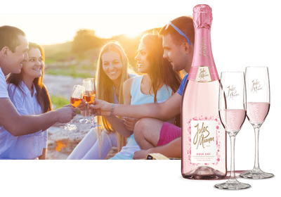 Im Vordergrund steht eine Flasche Rosé-Sekt, während fünf Freunde in der Abendsonne anstoßen und sich unterhalten.