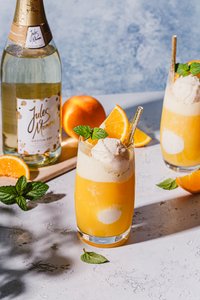 Zwei Gläser mit einem fruchtigen Cocktail, dekoriert mit einer Kugel Eis und frischen Minzblättern, auf einem hellen Tisch.