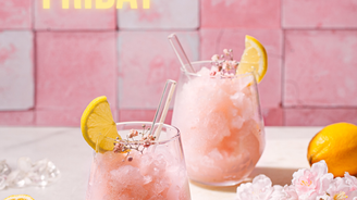 Zwei Gläser mit rosa gefrorenem Getränk, dekoriert mit Zitronenscheiben und kleinen Blüten, vor einer rosa Wand.
