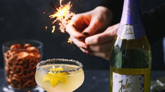 Eine Hand zündet eine Wunderkerze über einem Cocktail mit Sternenfrüchten, umgeben von Brezeln und einer Flasche Sekt.