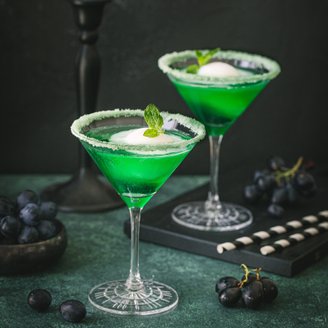 Zwei grüne Cocktails in eleganten Gläsern, dekoriert mit Minze und Zuckerrand, stehen auf einem dunklen Tisch.