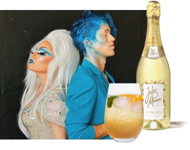 Ein kreatives Duo mit buntem Make-up und glitzernden Outfits steht Rücken an Rücken, während ein Getränk und Sekt im Fokus sind.