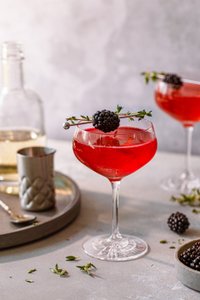 Ein eleganter Cocktail in einem Glas, garniert mit einer Brombeere und Thymian, auf einem hellen Tisch serviert.