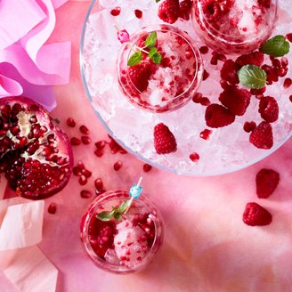 Drei Gläser mit gefrorenen Himbeeren und Granatapfelkernen auf Eis, umgeben von buntem Krepppapier.