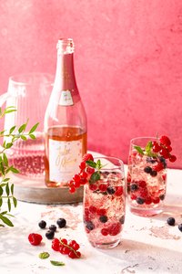 Auf einem Tisch stehen zwei Gläser mit prickelndem Rosé, dekoriert mit Beeren und Minze, während eine Flasche im Hintergrund sichtbar ist.