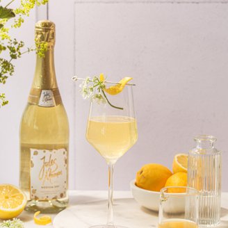 Ein elegantes Glas mit sprudelndem Getränk, garniert mit einer Zitronenschale und Blumen, steht auf einem marmorierten Tisch.