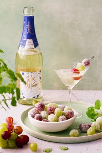 Ein stilvolles Martini-Glas mit sprudelndem Getränk und einer Garnitur aus bunten Trauben, neben einer Flasche Sekt platziert.