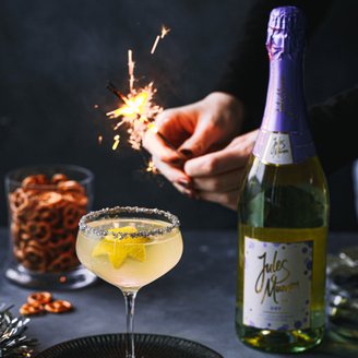 Eine Hand zündet eine Wunderkerze über einem Cocktail mit Sternenfrüchten, umgeben von Brezeln und einer Flasche Sekt.