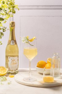 Ein elegantes Glas mit sprudelndem Getränk, garniert mit einer Zitronenschale und Blumen, steht auf einem marmorierten Tisch.